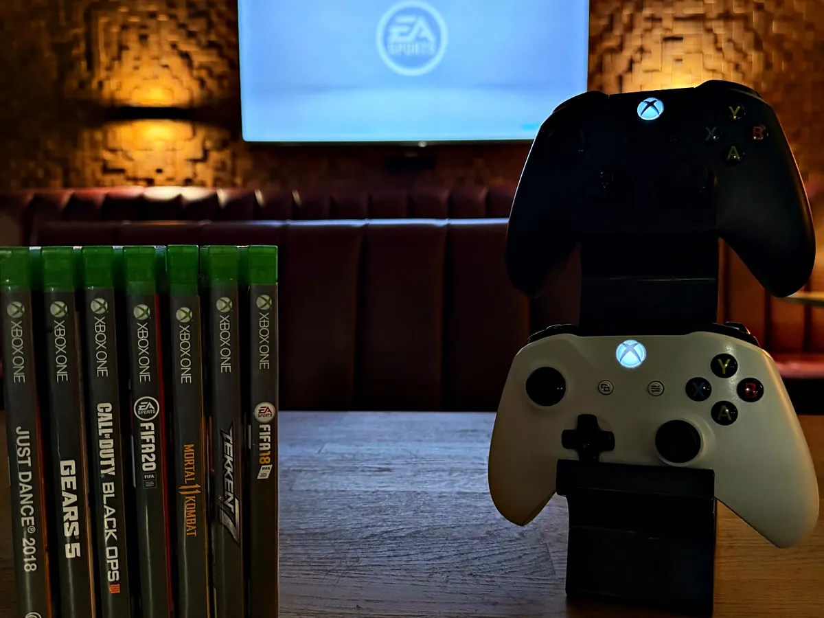 Dwa pady do Xboxa i gry na Xbox One na stoliku przed ekranem w Klubie Pictures